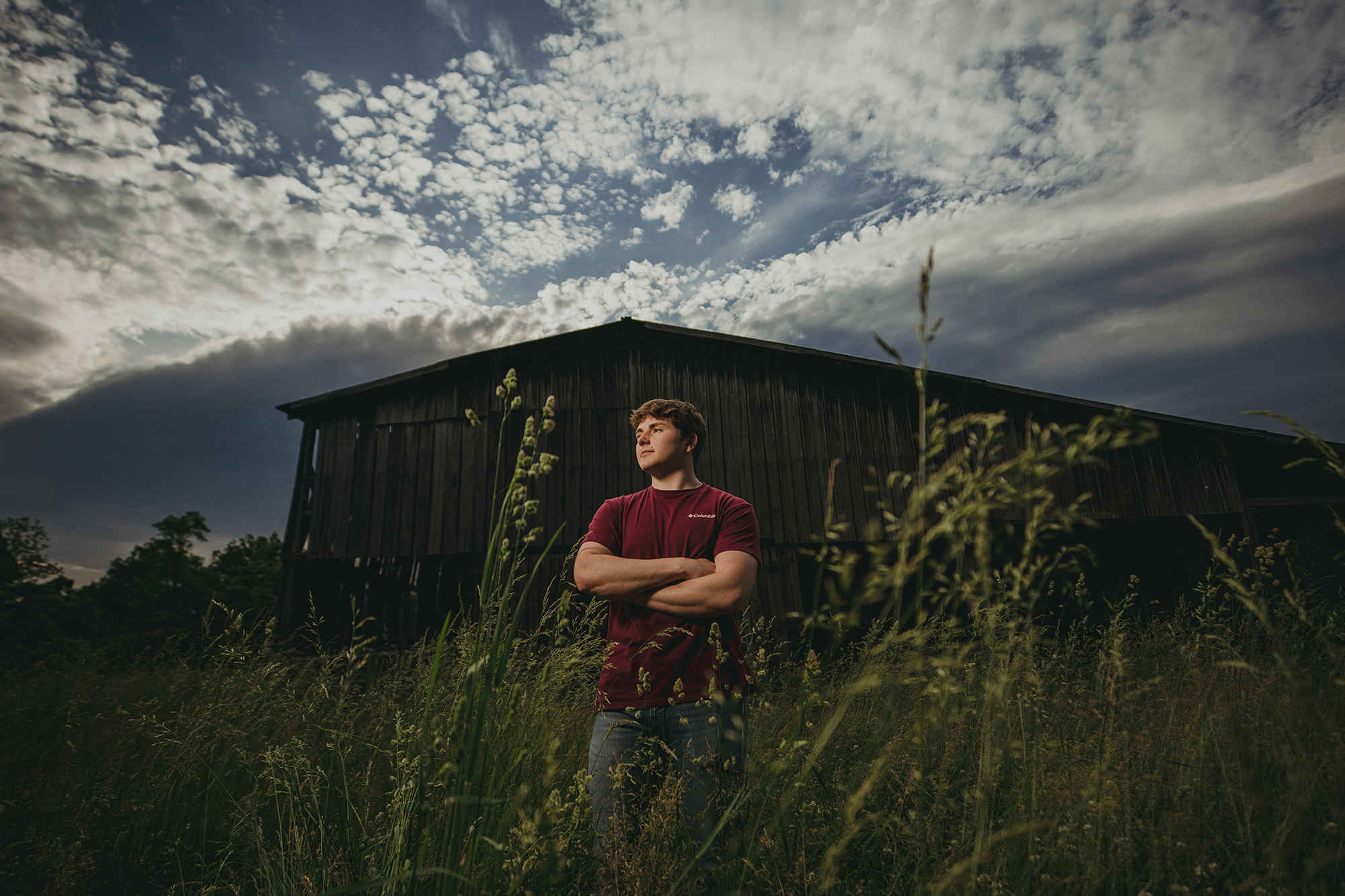 country senior boy with arms crossed in a farm field with tall grass and a barn in the background