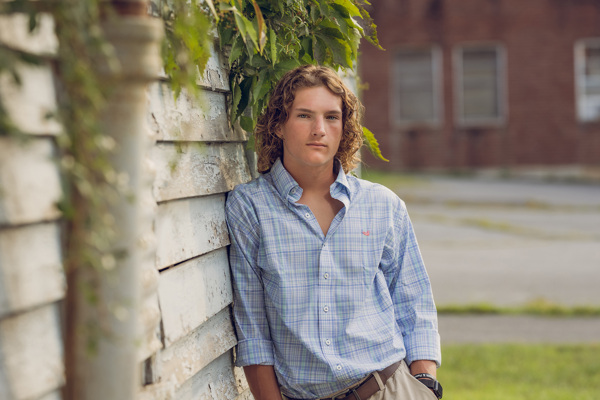 portrait of senior boy with long curly hair leaning up against an old house
